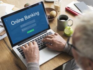 Überschulter Blick auf Hände eines Seniors auf einer Laptop-Tastatur mit Online-Banking Startseite auf dem Bildschirm