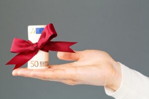 Ein gerollter 50 Euro-Schein mit roter Schleife drum steht auf einer ausgestreckten Handfläche<br />