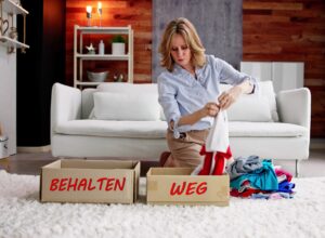 Eine dynamische Hausfrau kniet vor einem weißen Sofa auf einem weißen Teppich und sortiert Kleidung in zwei Pappkartons, mit jeweils der Aufschrift 