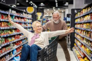 älterer Herr schiebt freudig ältere glückliche Dame in einem Einkaufswagen durch einen Supermarkt-Gang<br />