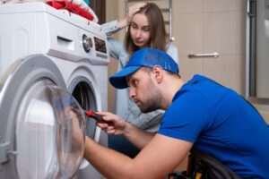 Handwerker im Blaumann schraubt an einer Waschmaschine, im Hintergrund eine genervte junge Frau