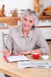 Dame Ü50 am Küchentisch, schaut glücklich beim Schreiben von Dokumenten, daneben ein Kaffee
