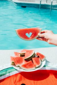 Teller mit Wassermelone vor einem Swimming Pool, eine Hand hält eine Wassermelonen-Scheibe hoch, aus der ein Herzchen ausgestanzt wurde